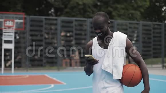 年轻的美籍黑人白人歌手拿着球看着他的智能手机边走边笑视频的预览图