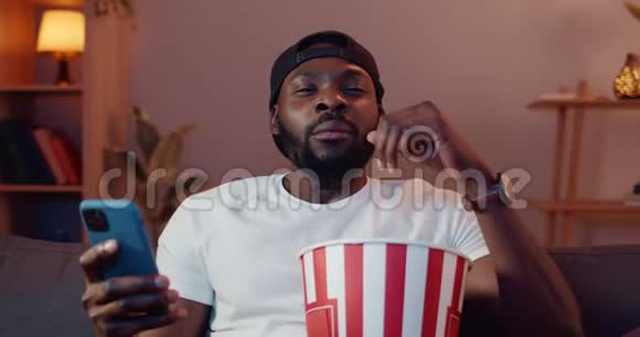 在电视机前拿着爆米花碗坐在沙发上年轻人一边打哈欠一边摇头无聊的非洲视频的预览图