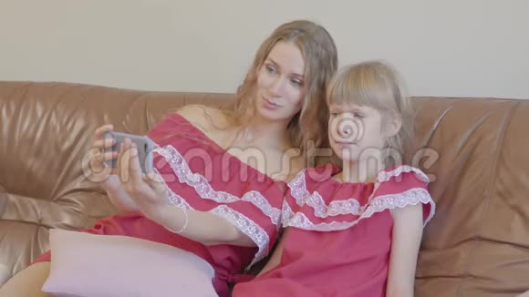 快乐的金发白种人妈妈和小女儿穿着同样的衣服坐在沙发上用手机自拍视频的预览图