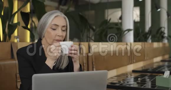 一位优雅的老女商人坐在桌子旁吩咐喝咖啡与笔记本电脑一起工作的成熟女性新手视频的预览图