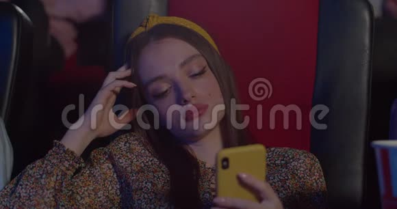 年轻女孩坐在电影院时使用她的智能手机美丽的千禧年女子厌倦了电影和视频的预览图