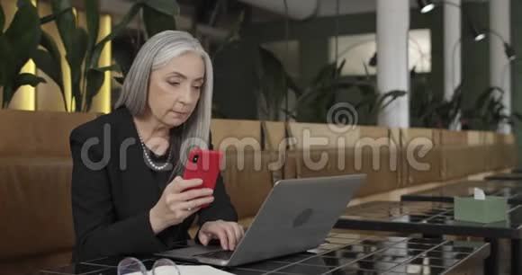 脾气暴躁的女企业主拿着智能手机在笔记本电脑上打字头发花白的老女商人视频的预览图