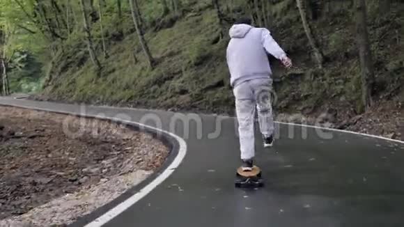 在蜿蜒的森林道路上乘坐灰色有帽衫的滑板游轮视频的预览图