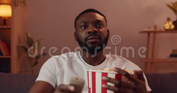 在电视上看有趣的电影一边吃爆米花一个满脸胡须的非洲男人年轻的帅哥坐在沙发上视频的预览图