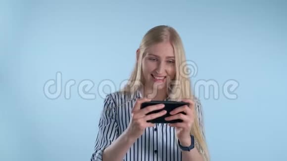 漂亮的金发女孩在蓝色背景下玩智能手机游戏视频的预览图