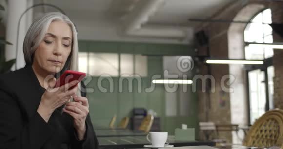 前景中年妇女企业主使用智能手机语音指挥官而坐在咖啡馆现代老太太视频的预览图