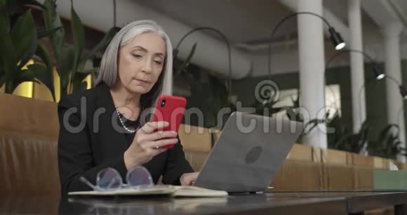 老妇人企业家看智能手机屏幕和在她的笔记本电脑上打字的正面视图成功的女商人视频的预览图