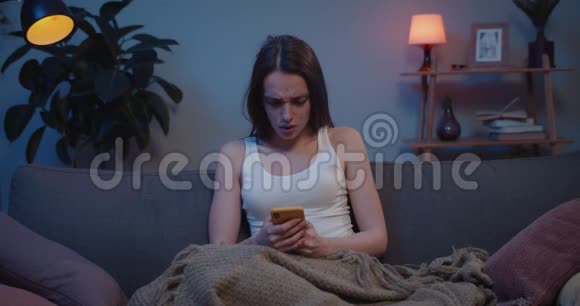 女孩坐在沙发上用智能手机和思考突发问题的正面视图年轻悲伤的女人捂脸视频的预览图