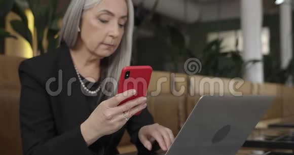 一个灰色头发的老妇人在咖啡馆里用智能手机和笔记本电脑工作成熟优雅的女商人视频的预览图