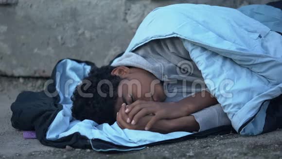 生病的黑人少年冻得瑟瑟发抖睡袋覆盖着躺在街上贫穷视频的预览图