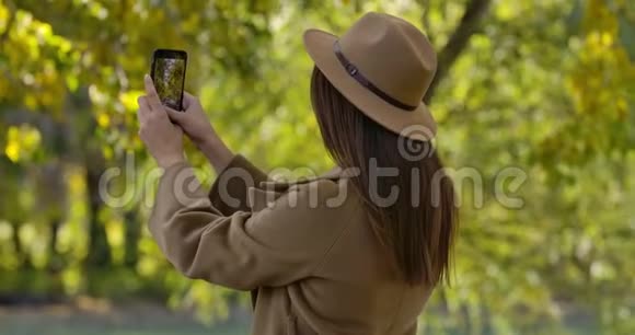 黑黑肤色的白种人女孩戴着棕色帽子和外套在她的智能手机上拍摄秋天公园的照片令人愉快视频的预览图