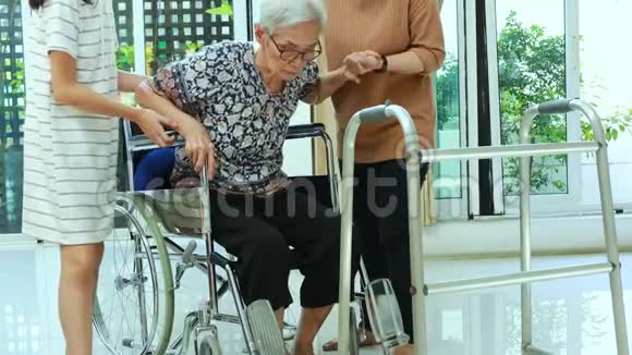 女性照顾者或护士支持帮助老年妇女在家里从轮椅上站起来老年母亲使用步行器练习WA视频的预览图