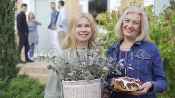 两个白种人成熟的女人微笑着拿着蛋糕和一桶花后台走廊上的公司视频的预览图