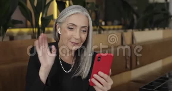 年长的漂亮女人穿着耳脚挥舞着智能手机相机视频对话成熟优雅视频的预览图