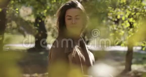 美丽的白种人女孩蓬松的发型和愉快的微笑站在秋天公园的黄叶后面很漂亮视频的预览图