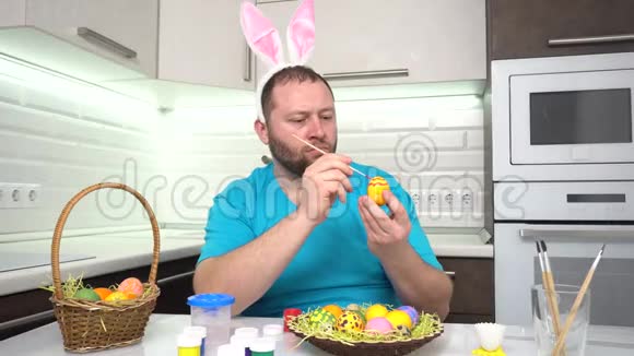 复活节快乐一个穿着兔子耳朵蓝色t恤的男人在厨房里画准备复活节的鸡蛋视频的预览图