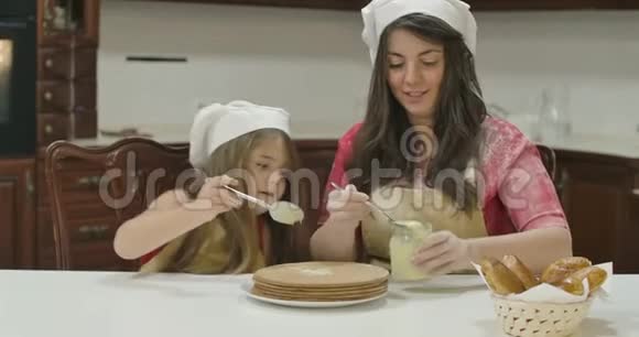 白种人的母亲和女儿在烘焙蛋糕上涂上炼乳戴厨师帽和围裙做饭的幸福家庭视频的预览图