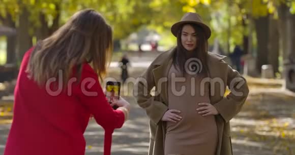可爱的高加索女人穿着优雅的棕色衣服和帽子在秋天公园的背景下为她的女朋友摆姿势视频的预览图