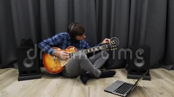 人类调谐电吉他戴耳机的人一边坐在客厅的地板上一边用笔记本调吉他年轻教授视频的预览图