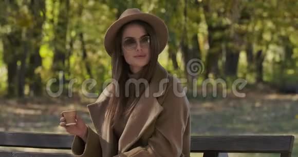 戴着墨镜和棕色帽子的白种人迷人的女士坐在长凳上喝着茶或咖啡微笑着视频的预览图
