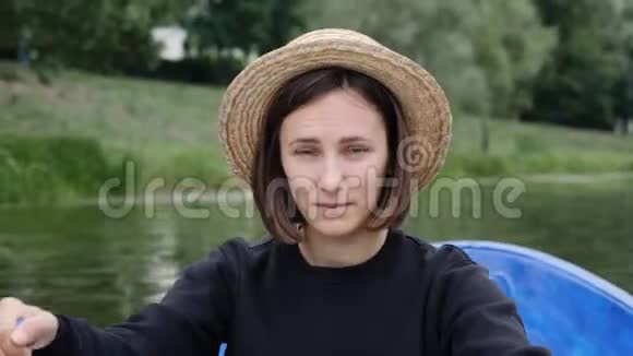 年轻的黑发女孩戴着草帽对着镜头微笑迷人的女孩在湖边的木船上航行在城市的船上飘浮的女孩视频的预览图