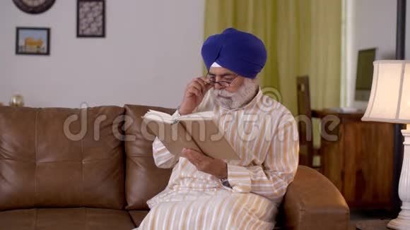 戴着蓝色头巾的锡克老人在他的客厅里的一盏灯旁看书退休生活视频的预览图