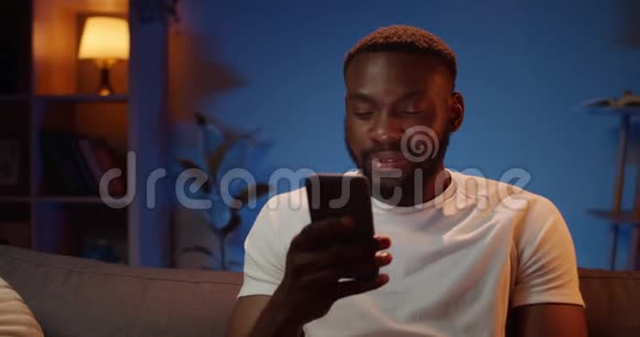 快乐的非洲男人坐在沙发上用智能手机与朋友交流年轻人的正面视野视频的预览图