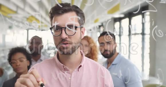 一名戴眼镜的男性办公室工作人员在玻璃板上写字并向团队成员解释概念帅哥特写视频的预览图
