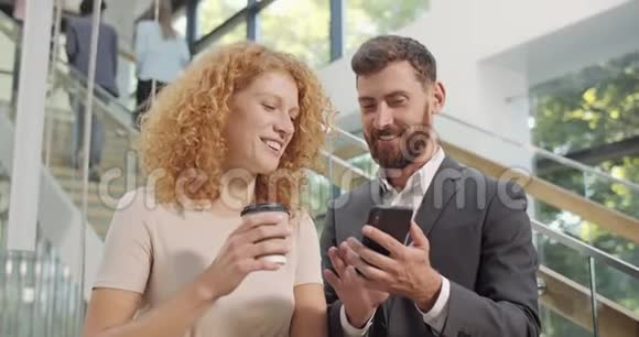 30多岁的英俊上班族向智能手机的女同事展示了有趣的照片站在大厅里的同事视频的预览图