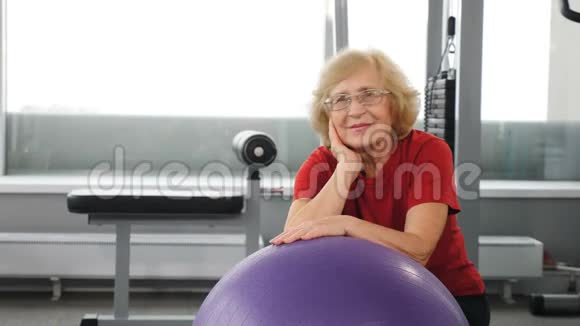 微笑的高级妇女休息后锻炼健身球在健身俱乐部里老太太靠在橡皮球上健康视频的预览图