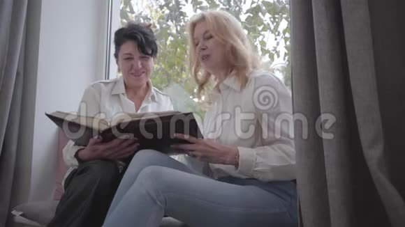 两个成熟的白人女人坐在窗台上看照片在家分享记忆的资深女性朋友视频的预览图
