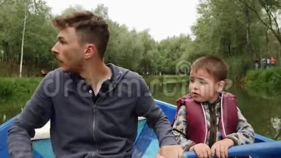年轻快乐的父亲驾驶木船儿子帮助他父亲和儿子在船上划船在我的绿色公园里互相交谈视频的预览图
