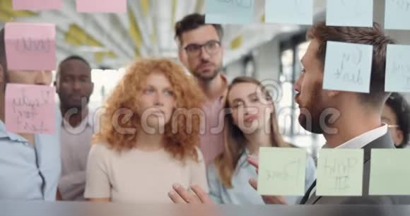 一群男工和女工在玻璃板附近用黏糊糊的音符进行热烈的讨论30岁男性商业教练视频的预览图