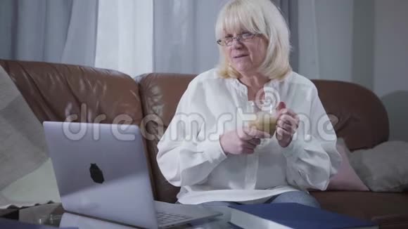 聪明的资深女性戴眼镜在笔记本电脑屏幕上看照片或照片喝咖啡白种人视频的预览图