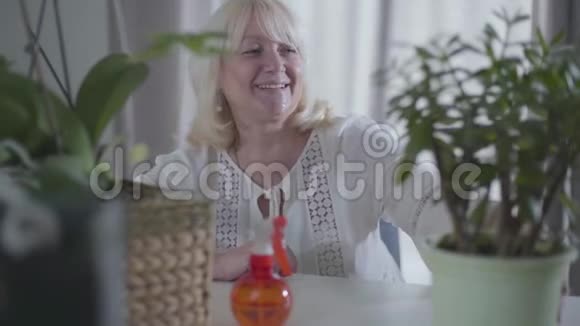 开朗的金发白种人退休者带着愉快的微笑和灰色的眼睛看着她的家乡植物和微笑肖像视频的预览图