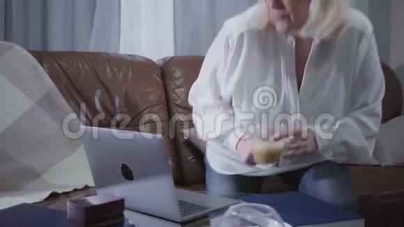 积极的高加索退休者坐在沙发上拿着咖啡杯看着笔记本电脑屏幕年长的金发女人视频的预览图