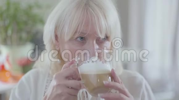 成熟的白种人金发女人的特写脸灰色的眼睛喝着咖啡看着远处孤独退休人员开支视频的预览图