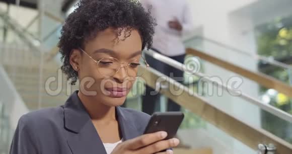 在现代办公大楼里用手机和微笑关闭戴眼镜的非洲女商人女办公室工作人员视频的预览图