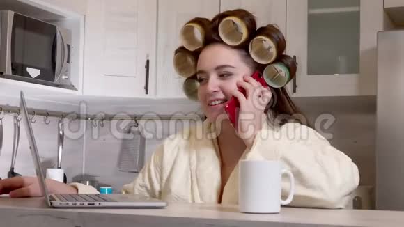 穿着浴袍头上戴着卷发器坐在厨房里用电话交谈视频的预览图