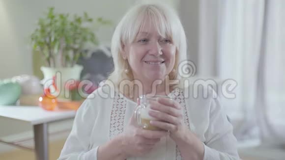 迷人的白种人高级妇女的肖像金发和灰色的眼睛喝咖啡和微笑快乐退休视频的预览图