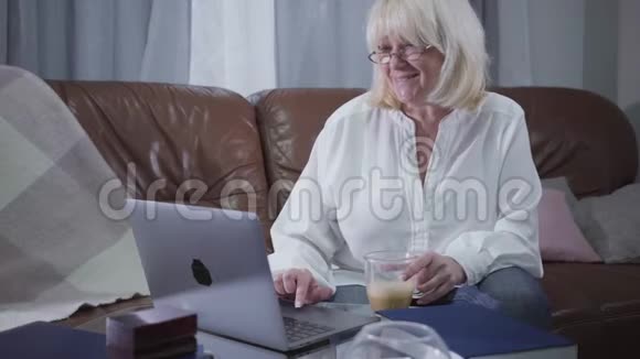 白种人芬梅尔退休者戴着眼镜坐在沙发上用咖啡杯和笔记本电脑的肖像白种人视频的预览图
