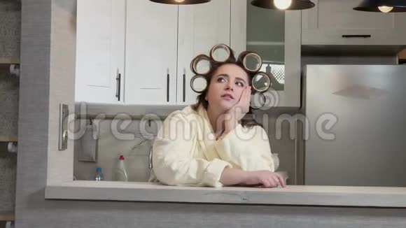 此外一个穿着浴袍的女孩在卷发器的头上独自坐在厨房里无聊视频的预览图