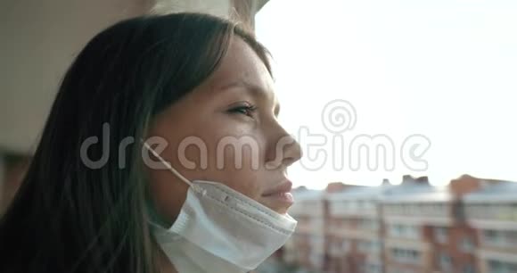一个戴着医用口罩的年轻女孩从窗口望向街上他公寓里女孩的自我隔离视频的预览图
