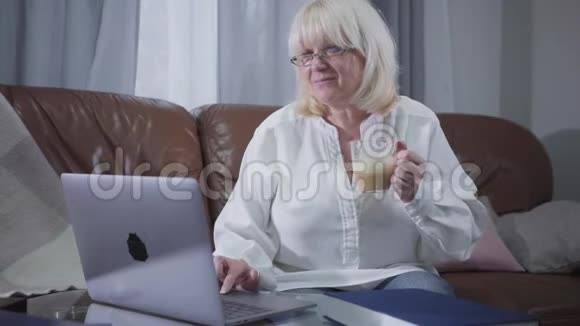 积极的白人戴眼镜的高级妇女坐在沙发上喝咖啡和上网金发成熟退休视频的预览图