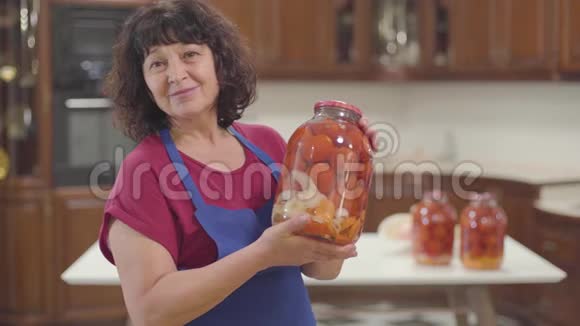 积极的白人祖母棕色的眼睛和黑色的卷发站在厨房里拿着一瓶番茄酱和视频的预览图