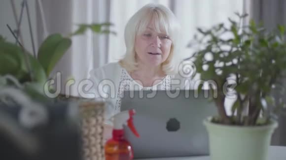漂亮的金发成熟的女人坐在家里的笔记本电脑后面她的园艺高加索退休人员坐着的肖像视频的预览图