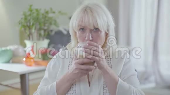 美丽的白种人成熟的女人金发和灰色的眼睛喝咖啡看着远方孤独退休人员开支视频的预览图