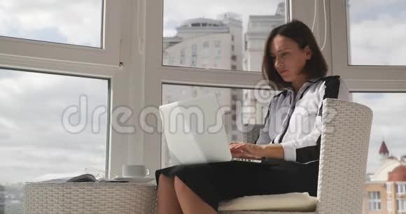 年轻漂亮的黑发商务女性坐在现代的白色椅子上喝着咖啡看着大城市视频的预览图