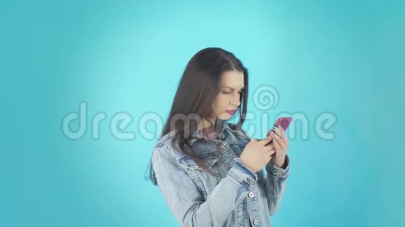 牛仔夹克里的年轻女子在蓝色背景下用智能手机写留言视频的预览图