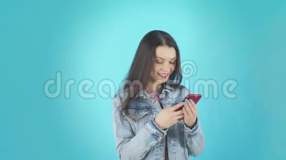 牛仔夹克里的年轻女子在蓝色背景下用智能手机写留言视频的预览图